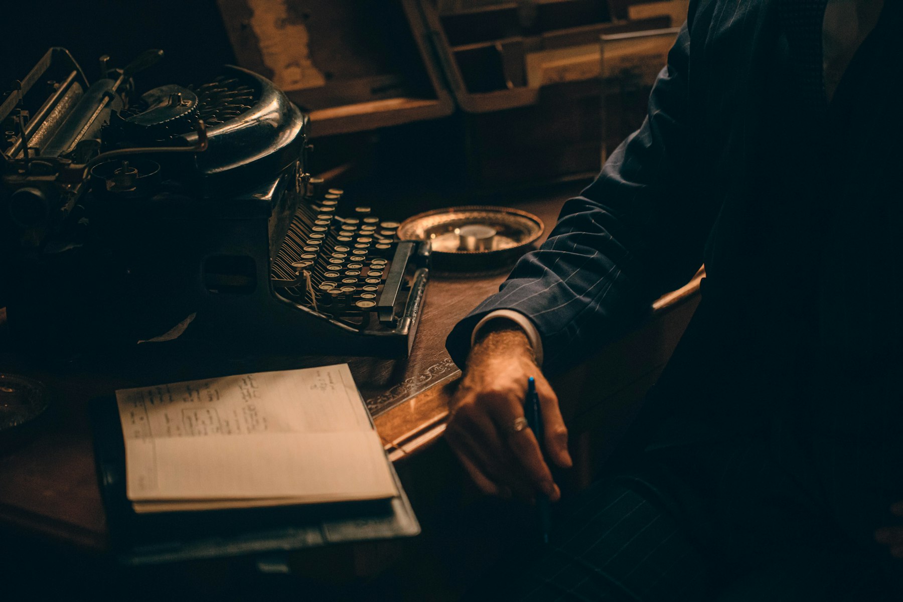 A writer with pen in hand beside a typewriter and an open notebook, in warm low light